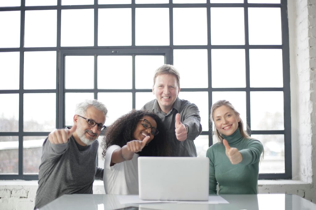 Happy multiracial team sitting at table with laptop and working on successful project while demonstrating thumb up gesture and smiling at camera